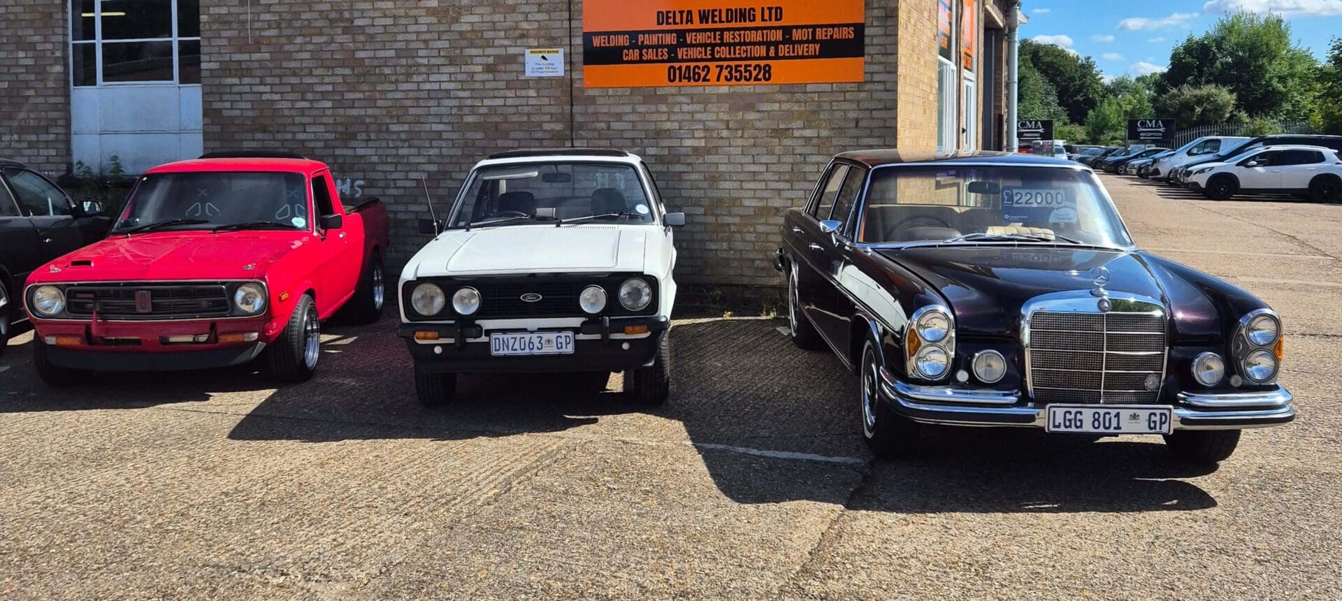 Two classic cars parked side by side on a sunny day outside a brick building.