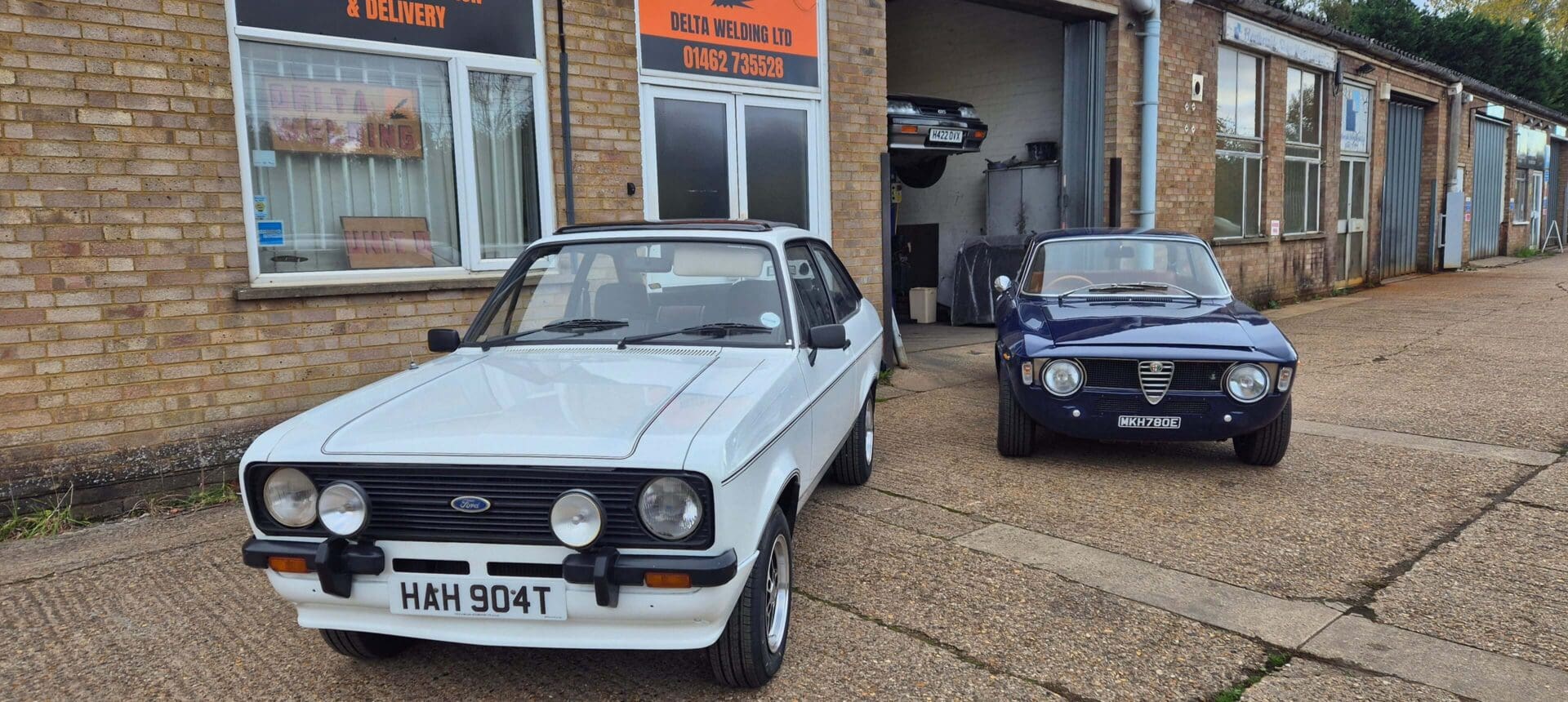 Two classic cars parked outside a workshop on a sunny day.