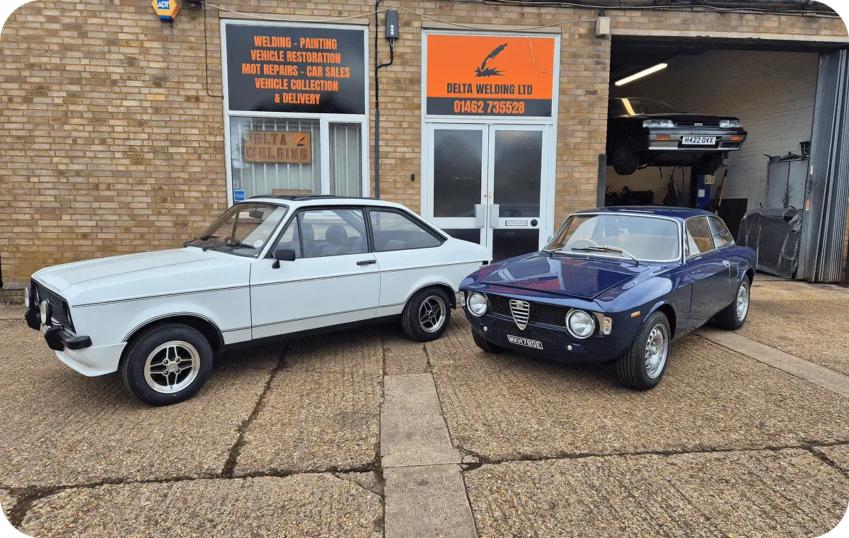 Two classic cars parked outside a vintage car workshop.